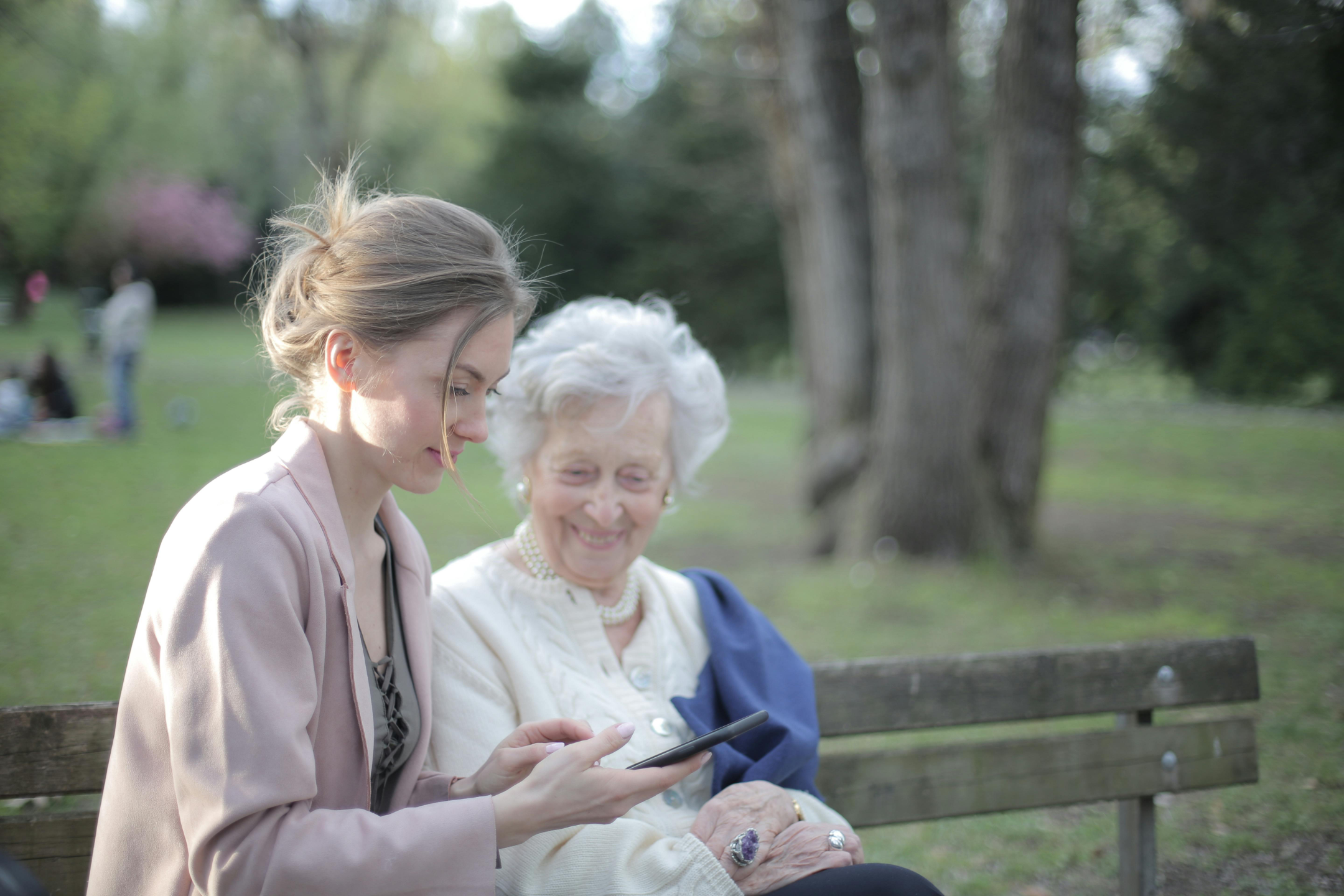 Eine Frau und eine Seniorin sitzen auf einer Bank und schauen auf ein Smartphone